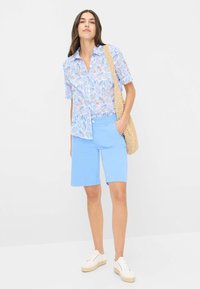 Kurzärmliges Hemd in Hellblau mit mehrfarbigen Blattmustern, kombiniert mit hellblauen Shorts und weißen Sneakers.