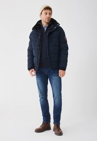 Puhasto tamno plava jakna s kapuljačom, bočnim džepovima i zatvaračem na munje. Kombinirano s plavim džemperom, trapericama i smeđim čizmama.