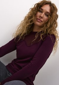 Top bordeaux a maniche lunghe con pannello trasparente e scollo ondulato, realizzato in un tessuto morbido e elasticizzato. Il modello indossa pantaloni grigi.