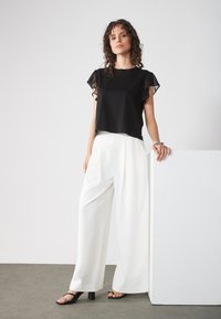 Haut noir à manches courtes avec des manches en voile à volants, associé à un pantalon blanc à jambes larges. Le modèle porte des sandales noires à talons. Conception simple.