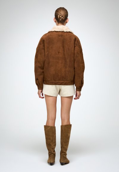 Bruine suède jas met een fluffy crèmekleurige kraag, oversized pasvorm en lange mouwen, gecombineerd met beige shorts en kniehoge bruine suède laarzen.