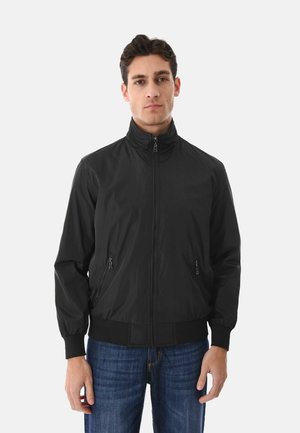 Giovane uomo che indossa una giacca nera con cerniera e collo alto, con tasche laterali con cerniera, abbinata a jeans blu, in piedi davanti a uno sfondo bianco.