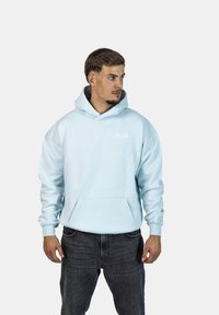 Felpa blu chiaro realizzata in materiale morbido, dotata di tasca a marsupio e cappuccio con cordino. Presenta il logo "CRYSTAL" in bianco. Indossata con jeans scuri.