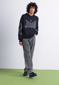 Sort hættetrøje med "JORDAN" tekst, parret med grå mønstrede jeans og sorte sneakers med blå accenter. Modellen står på grønnt gulv.