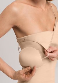 Naga, w kolorze nude, odzież z regulowanymi ramiączkami; ręka trzymająca gładkie, okrągłe wsparcie, wykonane z miękkiego, elastycznego materiału.