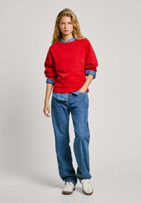 Felpa rossa con vestibilità comoda, maniche lunghe e dettagli a coste sui polsini. Indossata sopra una camicia di denim chiaro e abbinata a jeans blu larghi e sneakers.