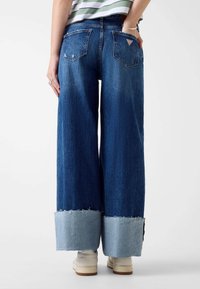 Jeans a gamba larga di colore blu con un orlo sbiadito di un blu chiaro, dotati di tasche posteriori e un piccolo logo sulla tasca destra.