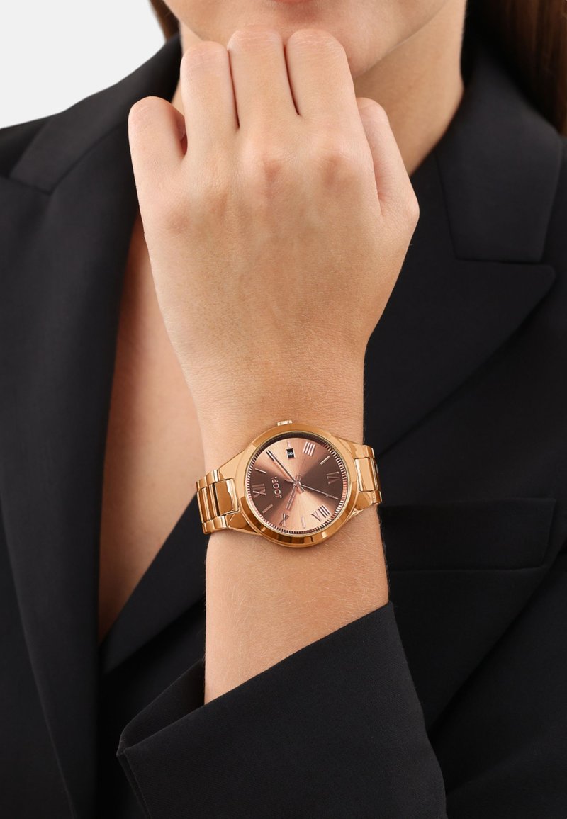 Roze gouden horloge met een ronde wijzerplaat, Romeinse cijfers als markeringen en een metallic schakelhalsband, gedragen aan een hand tegen een zwarte blazer.