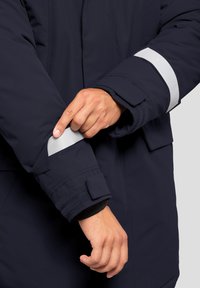 Giacca puffer blu navy con strisce riflettenti argentate sui polsini. Presenta chiusure in velcro e una morbida fodera interna per maggiore comfort.