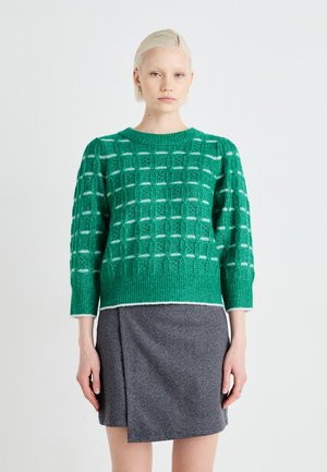 Green knitted sweater with a textured, patterned design, featuring three-quarter sleeves and white accents at the collar and hem. Grey skirt.