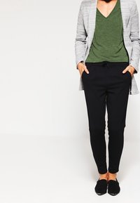 Grauer Blazer über einem grünen V-Ausschnitt-Top und schwarzen, taillierten Hosen. Das Outfit hat eine lockere Passform, mit einem Kordelzugbund und glatter Textur.