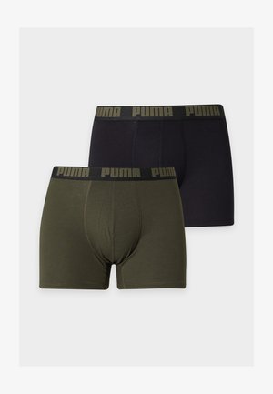 Szczupły mężczyzna o ciemnej karnacji stoi boso, ubrany w ciemnozielone bokserki marki Puma, ręka spoczywa na dolnej części pleców, neutralne szare tło.