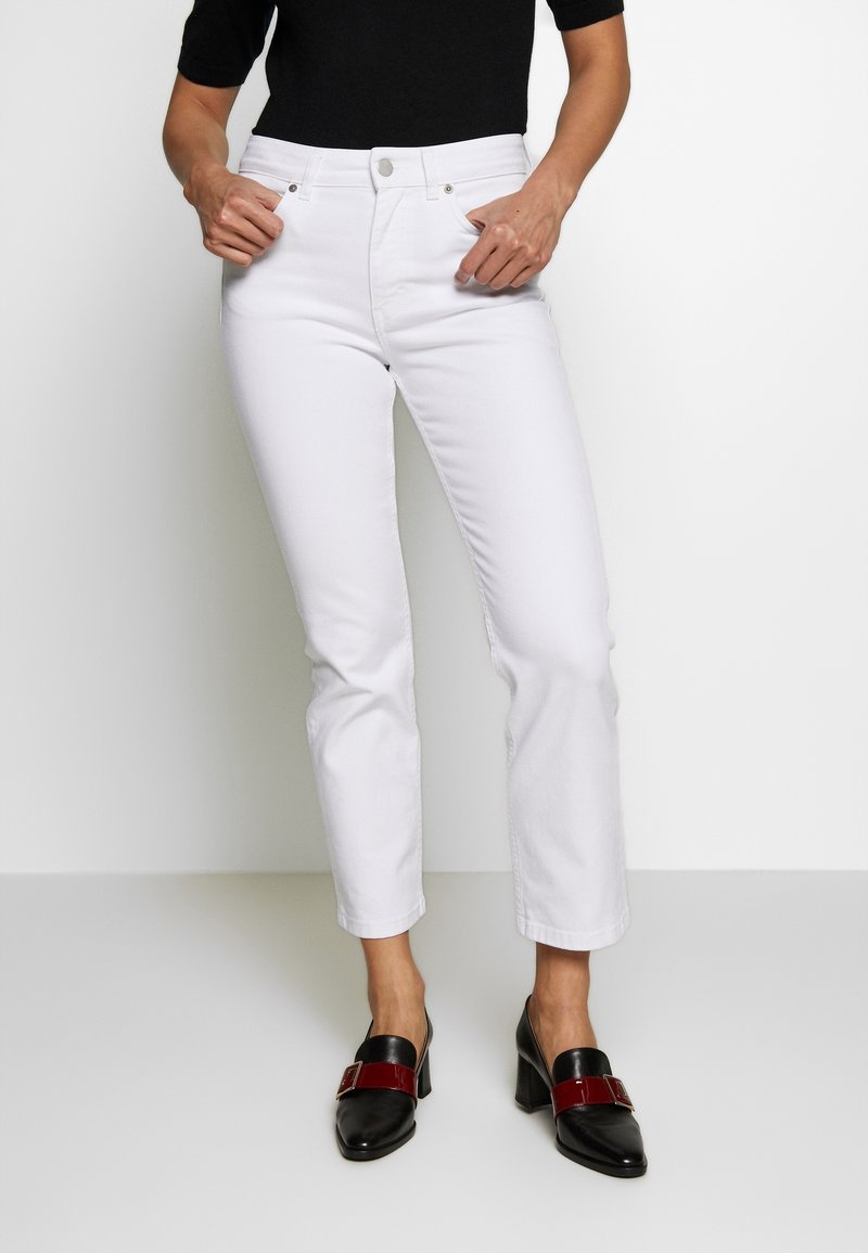 Jeans blancos de pierna recta con cintura alta y diseño clásico de cinco bolsillos. Combinados con tacones negros que presentan un detalle de tira roja.