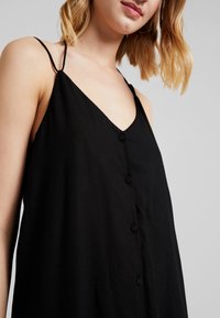 Robe noire sans manches avec un décolleté en V, des boutons sur le devant et des bretelles fines. Le tissu semble léger avec une texture lisse.
