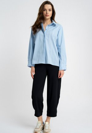 Kobieta ubrana w jasnoniebieską koszulę na guziki w paski, czarne spodnie za kostkę oraz beżowe trampki, stojąca na tle jednolitego białego tła.