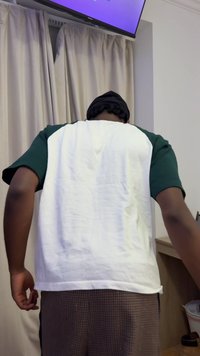 Kurzärmeliges Hemd aus weißem und grünem Stoff, kombiniert mit braunen karierten Hosen. Der Hintergrund zeigt Vorhänge und eine niedrige Holzoberfläche.