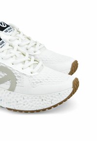 Witte sneakers met een gestructureerde gebreide bovenkant, voorzien van een groot grijs logo, reflecterende accenten en een gespikkelde witte tussenzool met een bruine rubberen buitenzool.