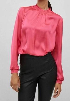 Femme portant un chemisier en satin rose à manches longues avec un col et des poignets froncés, assorti à un pantalon noir taille haute.