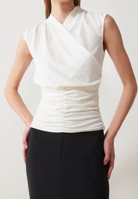 Top blanco sin mangas con diseño envolvente y textura fruncida en la cintura, combinado con una falda negra sólida. Tela suave, detalles mínimos.