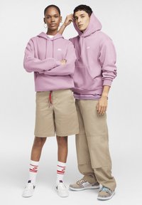 Lila jopa s kapuco z belimi logotipi, bež kratke hlače in nogavice Nike; superge v sivi in beli barvi. Stojijo pred enostavnim ozadjem.