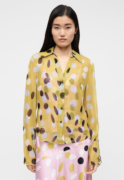 Priehľadná žltá blúzka s viacfarebnými kruhovými vzormi, klasickým golierom a čiernymi gombíkmi, v páre s ružovou sukňou s podobnými bodkami.