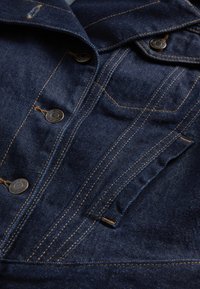 Tume teksatagi nööpkinnitustega ja kontrastse oranži õmblusega. Omab klassikalist kraega kaelust ja rinnataskuid peene detailiseerimisega.