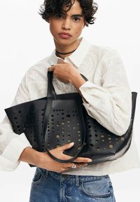 Schwarze, perforierte Tragetasche mit runden Ausschnitten, flachen Griffen und einer strukturierten Form. Aus Leder hergestellt, das eine glatte Textur aufweist.