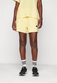 Žlté športové šortky s hladkou textúrou, s čiernym logom Adidas. V kombinácii s čiernymi teniskami a bielymi pruhovanými ponožkami.