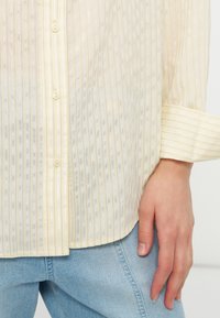 Ανοιχτό κίτρινο πουκάμισο με κουμπιά και κάθετες ρίγες, μακριά μανίκια με μανσέτες, φορεμένο πάνω από ανοιχτόχρωμο μπλε τζιν, με το χέρι χαλαρό στο πλάι.