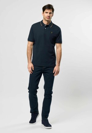 Bărbat stând, îmbrăcat într-un tricou polo bleumarin închis cu margini bej, blugi închiși și adidași bleumarin, pe un fundal simplu, deschis la culoare.