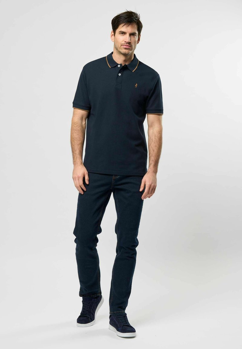 Bărbat stând, îmbrăcat într-un tricou polo bleumarin închis cu margini bej, blugi închiși și adidași bleumarin, pe un fundal simplu, deschis la culoare.