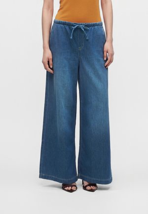 Femeie purtând pantaloni din denim albaștri, cu croială largă și talie cu șnur, și pantofi negri cu vârf deschis, stând cu un fundal alb.