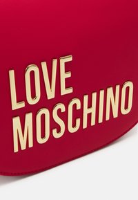 Červená syntetická kabelka s výrazným zlatým nápisem "LOVE MOSCHINO." Hladká textura s zaobleným tvarem a viditelnými švy.