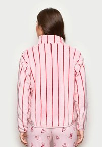 Kobieta z długimi brązowymi włosami, ubrana w różową polarową kurtkę w pionowe czerwone paski oraz różowe spodnie z wzorem w czerwone cukrowe serca.