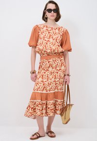 Rotes und cremefarbenes Blumenkleid mit Puffärmeln, tailliertem Schnitt und gestuftem Saum, kombiniert mit einer Strohtasche und braunen Sandalen.