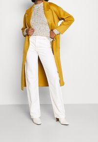 Persoon in een mosterdgele jas, lichtgrijs gemêleerd truitje, witte hoge broek en witte hakkenlaarsjes, staand met handen op de heupen.