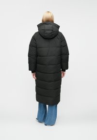 Hosszú fekete puffertakó kapucnival, steppelt részekkel és egyenes szabással. A modellen széles szárú farmernadrág van.