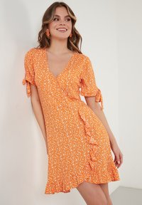 Oranje bloemenprintjurk met korte mouwen, V-hals en ruches aan de onderrand. Kenmerkt strikdetails op de mouwen en een lichtgewichtig stof.