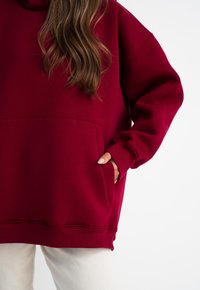 Felpa con cappuccio bordeaux, dotata di una grande tasca frontale, polsini a coste e vestibilità comoda. Tessuto morbido al tatto con leggero elasticità.