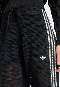 Čierne teplákové nohavice z priedušnej sieťoviny s bielym vzorom troch prúžkov, elastickým pásom a nastaviteľnou šnúrkou. Pri vrecku je malá biela logo.