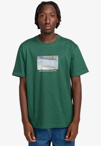 Grøn bomulds t-shirt med et rektangulært grafisk design, der har teksten "Hilsner fra Barcelona". Rund halsudskæring, korte ærmer, afslappet pasform.