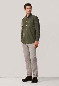 Camisa de manga larga verde oliva con cuello abotonado, bolsillo en el lado izquierdo del pecho, combinada con pantalones gris claro y zapatos oscuros.