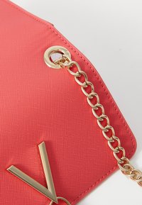 Borsa corallo brillante con una finitura testurizzata, caratterizzata da una tracolla in oro e un accento metallico dorato a forma di V.
