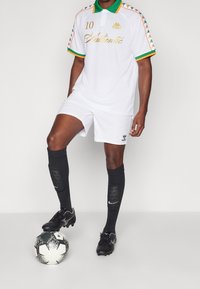 Bílý fotbalový dres se zelenými a oranžovými akcenty, zahrnuje košilku s límečkem a šortky, v kombinaci s černými ponožkami a kopačkami, stojící nad fotbalovým míčem.