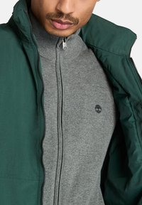 Grüne Zip-Jacke über einem grauen Reißverschluss-Pullover. Die Jacke hat eine glatte Textur, während der Pullover ein gestricktes Muster und ein kleines Logo aufweist.