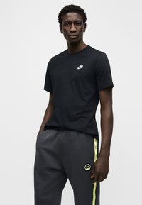 Maglietta Nike nera con piccolo logo bianco; abbinata a pantaloni grigio scuro con strisce gialle e patch con logo sulla coscia.