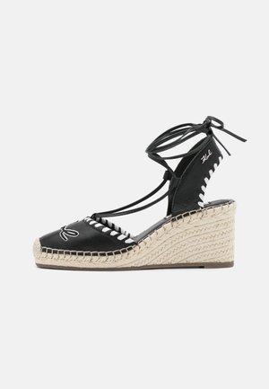 Sort kileespadrille med ankelviklelisser, hvide syninger og flettet halm hæl på en ensartet lys baggrund.