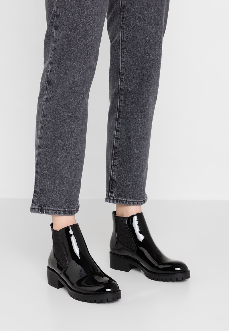 Jambes portant un jean gris foncé et des bottines en cuir verni noir brillant à talons épais sur un sol gris clair.