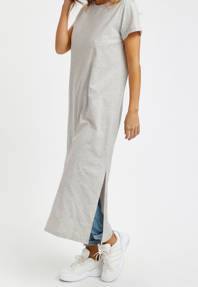 Femme portant une robe maxi gris clair à manches courtes avec fente latérale sur un jean bleu et des baskets blanches sur fond blanc uni.