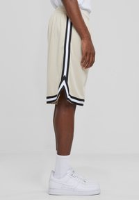 Beige sportshorts med mesh-struktur, der har sorte og hvide side-striber og en delt kant. Der er sat sammen med hvide sneakers og sokker.
